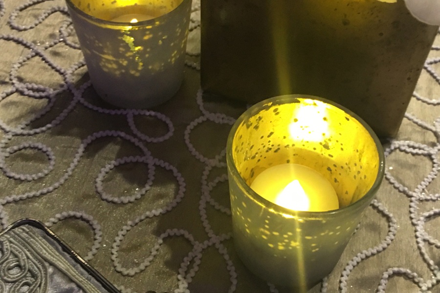 The flame of a candle burns. The candle is in a translucent container, set on a brocade tablecloth.