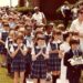 Children in school uniforms walk together with hands folded in a May Procession to honor the Blessed Mother