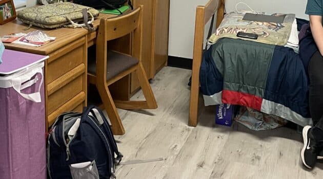 A college dorm room with a backpack, desk, part of a bed