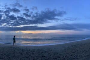 Wide shot of a sunrise on the beach, with a figure standing toward the left watching the light come into the sky
