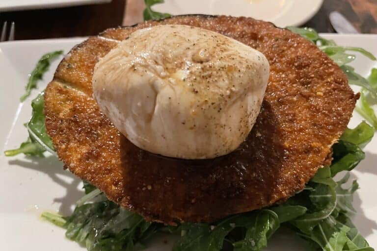 Caprese salad on a plate with a ball of mozzarella on a piece of fried eggplant atop greens
