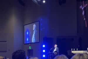 Fr. Greg Boyle, S.J., speaks from a podium with a crucifix hanging on one side and his image displayed on a video screen on the other side