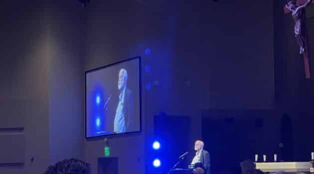Fr. Greg Boyle, S.J., speaks from a podium with a crucifix hanging on one side and his image displayed on a video screen on the other side