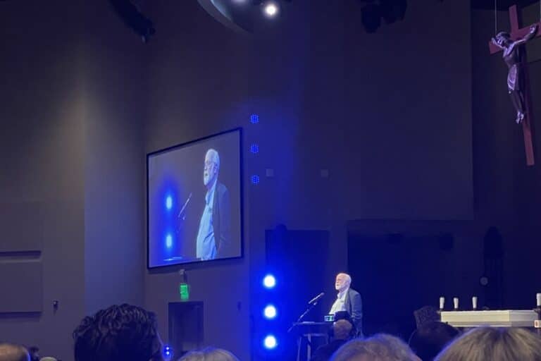 Fr. Greg Boyle, S.J., speaks from a podium with a crucifix hanging on one side and his image displayed on a video screen on the other side