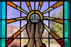 Stained glass window shows hands lifting a host with IHS on it