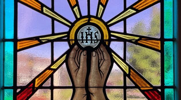 Stained glass window shows hands lifting a host with IHS on it