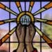 Stained glass window shows hands lifting a host with IHS on it