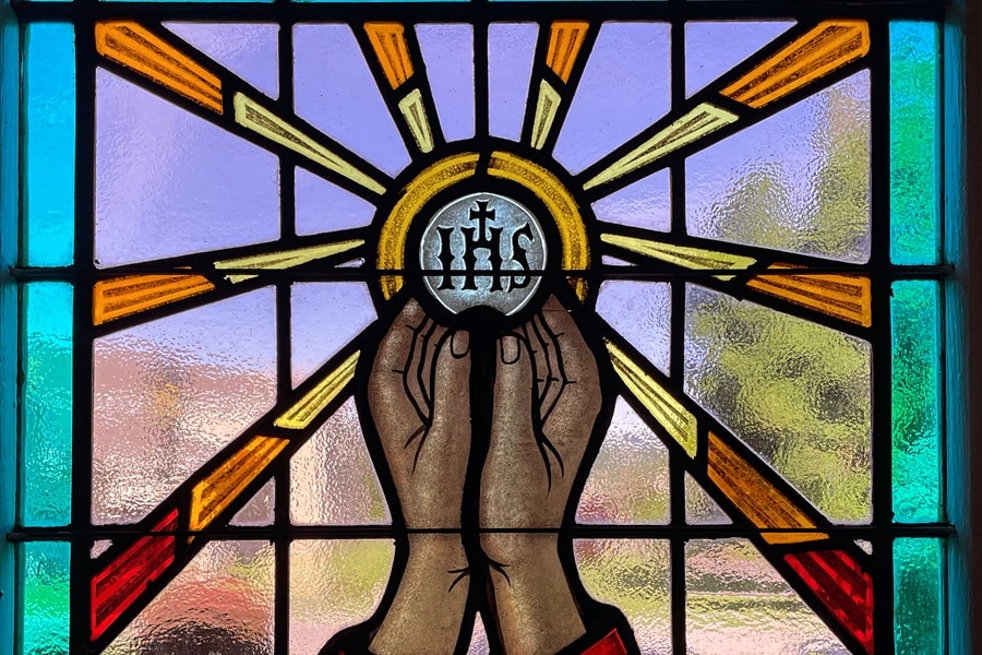 Stained glass window shows hands lifting a host with IHS on it
